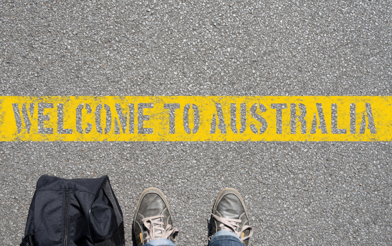 Feet and a bag standing next a welcome to Australia written on a yellow line in the pavement