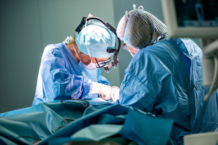 Surgeon and his assistant performing cosmetic surgery in hospital operating room. Surgeon in mask wearing loupes during medical procedure.