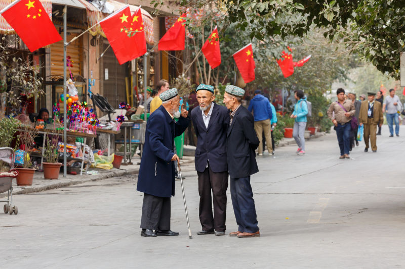 Three elderly, Uyghur men having a conversation. In the back Chinese flags.