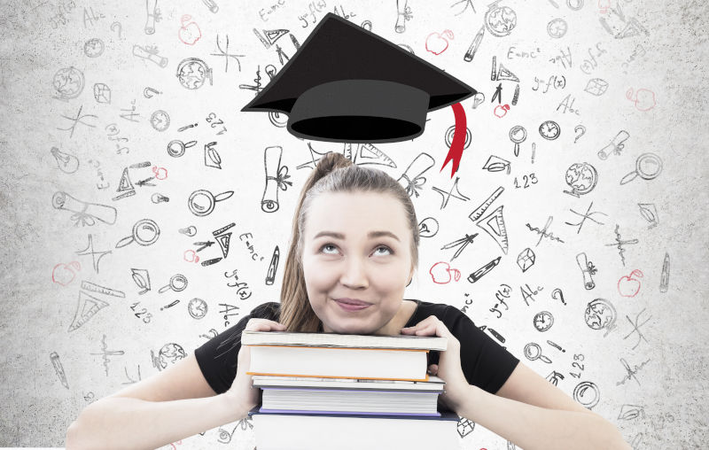 Woman is sitting at a table with her head on a pile of books. Concrete wall background with education icons and a graduation hat above her.