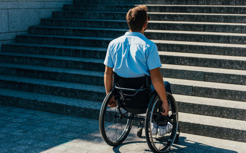 Back view of man using wheelchair on street and looking at stairs without ramp