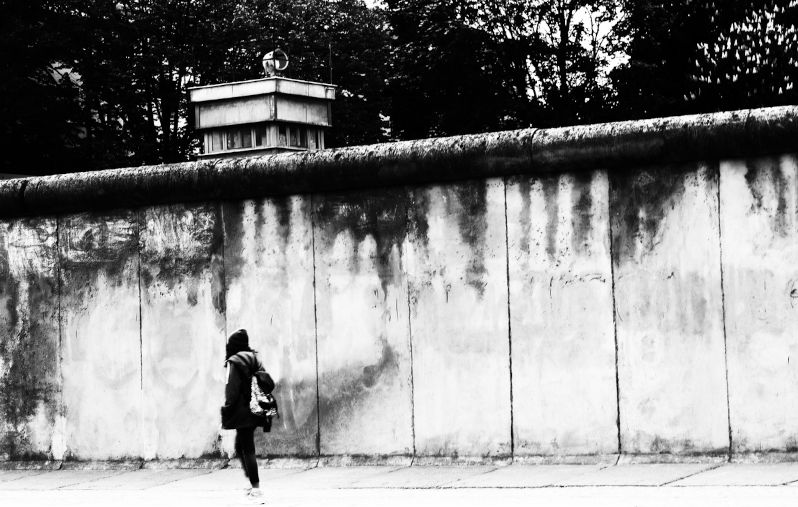 Black / white photograph of the Berlin Wall. A young woman walks carelessly along the wall. Behind the Berlin Wall can be seen, a former watchtower. Reminder of the division of Berlin. Former border fortification in Berlin.