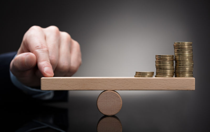 Finger Balancing Stacked Coins On Seesaw Against Grey Background.