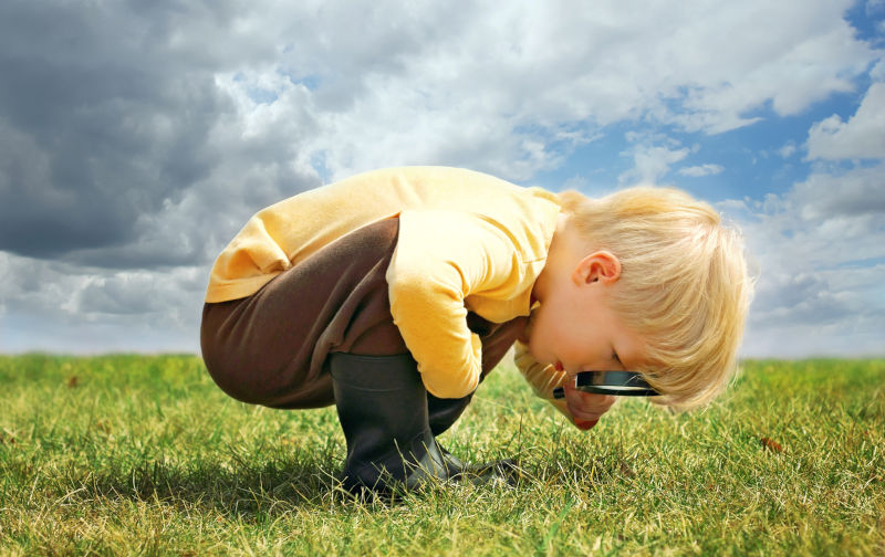 A little boy is exploring nature outside by looking at grass through a magnifying glass.