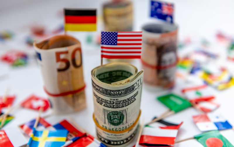 Rolled bundles of major currencies and country flags with the US Dollar and American flag in front. Dollar hegemony concept.