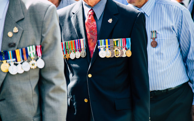 A formed body of old military veterans march past the crowd during the Cooroy-Pomona ANZAC Day parade. The man in the foreground proudly wears the golden Order of Australia Medal (OAM) along with campaign medals from The Vietnam War, the Infantry Combat Badge (ICB) and general Defence service medals. The man marching behind him wears his personal medals on his right breast and World War 2 service medals on his left side.