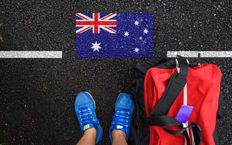 A man with a shoes and travel bag is standing on asphalt next to flag of Australia and border.